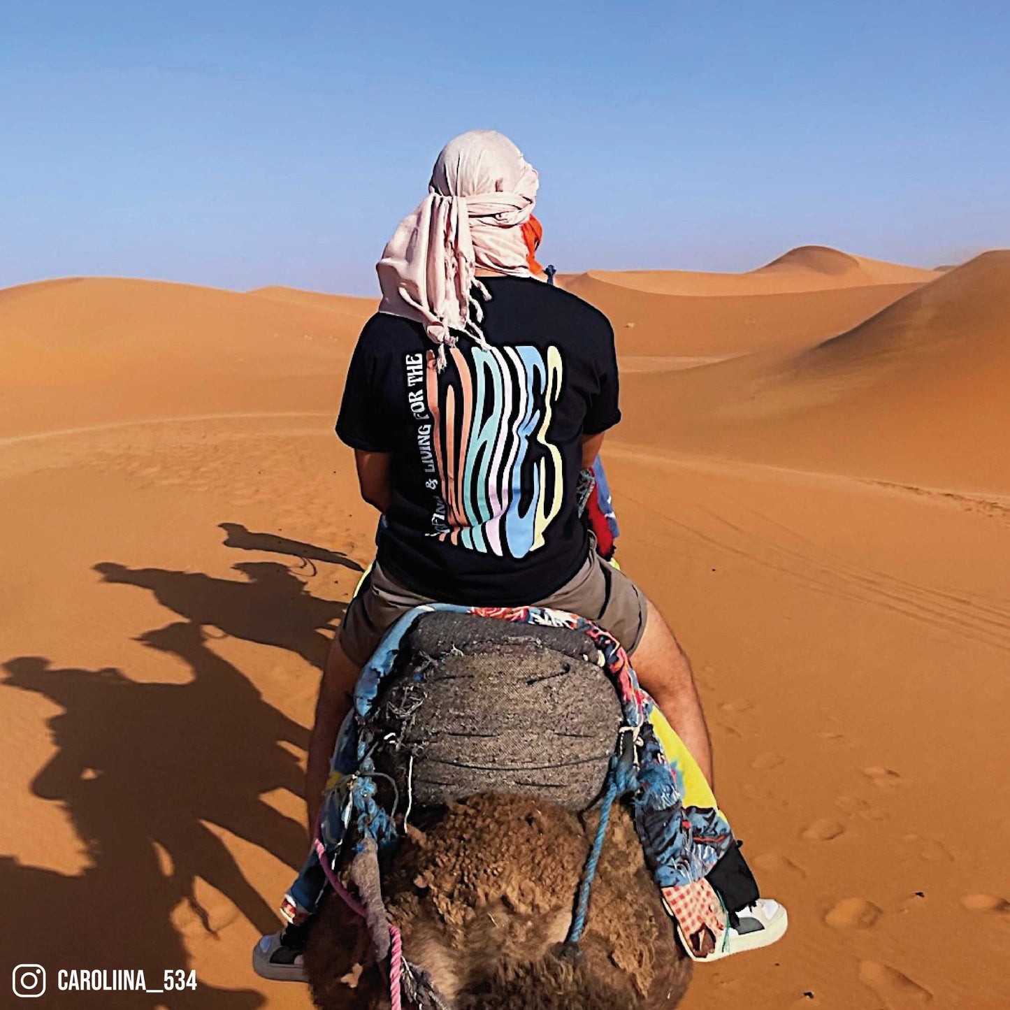 Photographie d'un homme sur un dromadaire dans le désert portant un tshirt noir orcaz designs en coton avec un design typographique coloré dans le dos