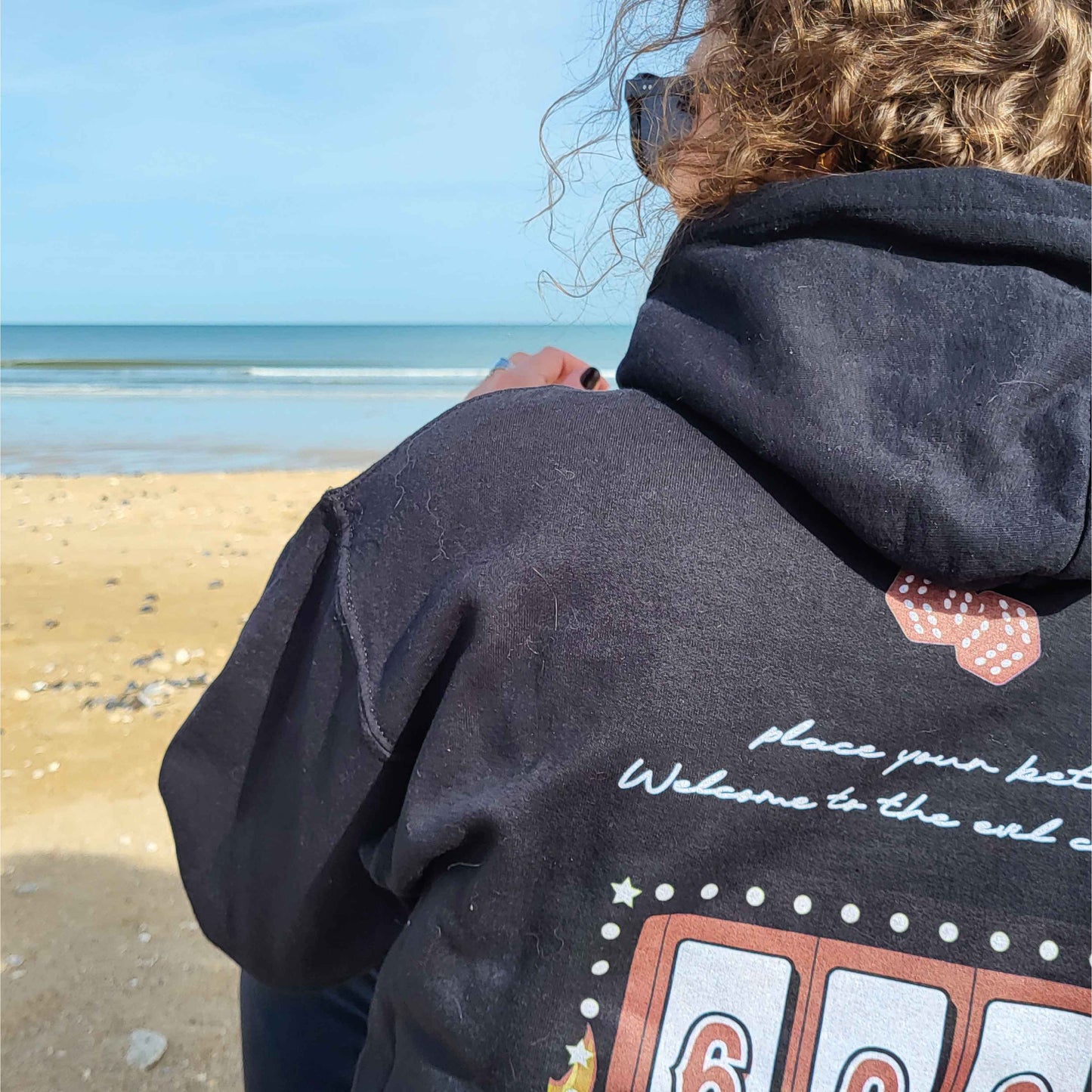 Photographie d'un Hoodie noir imprimé dans le dos d'un design de machine à sous de casino version enfer 666 porté par une femme sur la plage