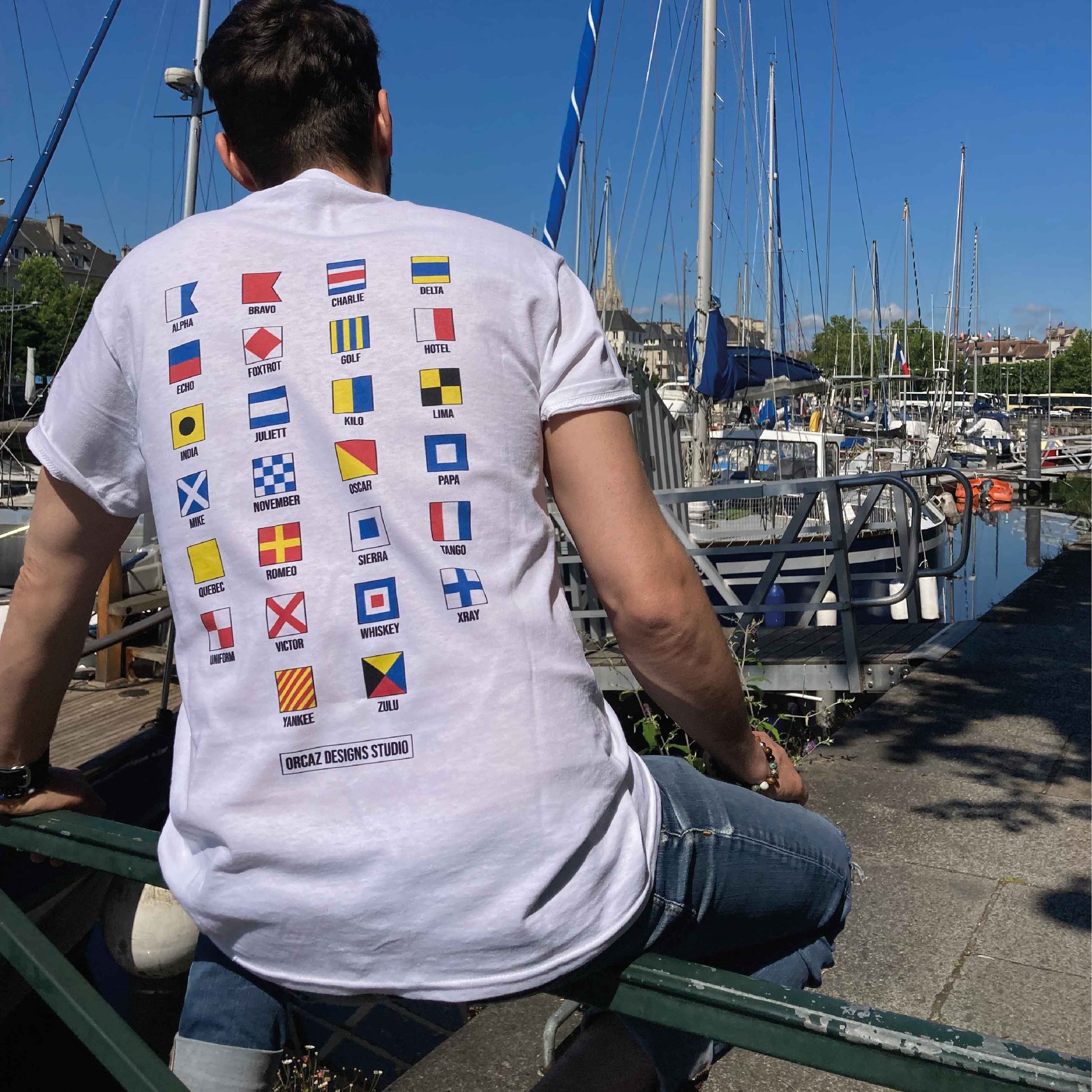 Photographie d'un tshirt blanc porté par un jeune homme avec l'alphabet maritime imprimé dans le dos et les voiliers du port de Caen en fond 