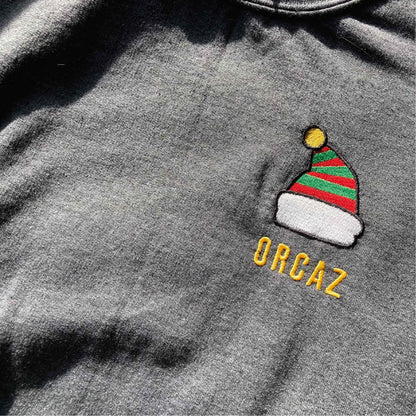 Photographie d'un sweat gris avec une broderie d'un bonnet d'Elfe du père Noël en couleurs avec la marque Orcaz brodée en jaune en dessous